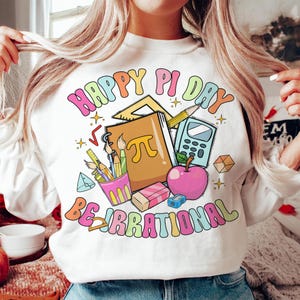 May include: White sweatshirt with a colorful graphic design that says "Happy Pi Day Be Irrational". The graphic includes a notebook, pencils, a calculator, a protractor, and a red apple.