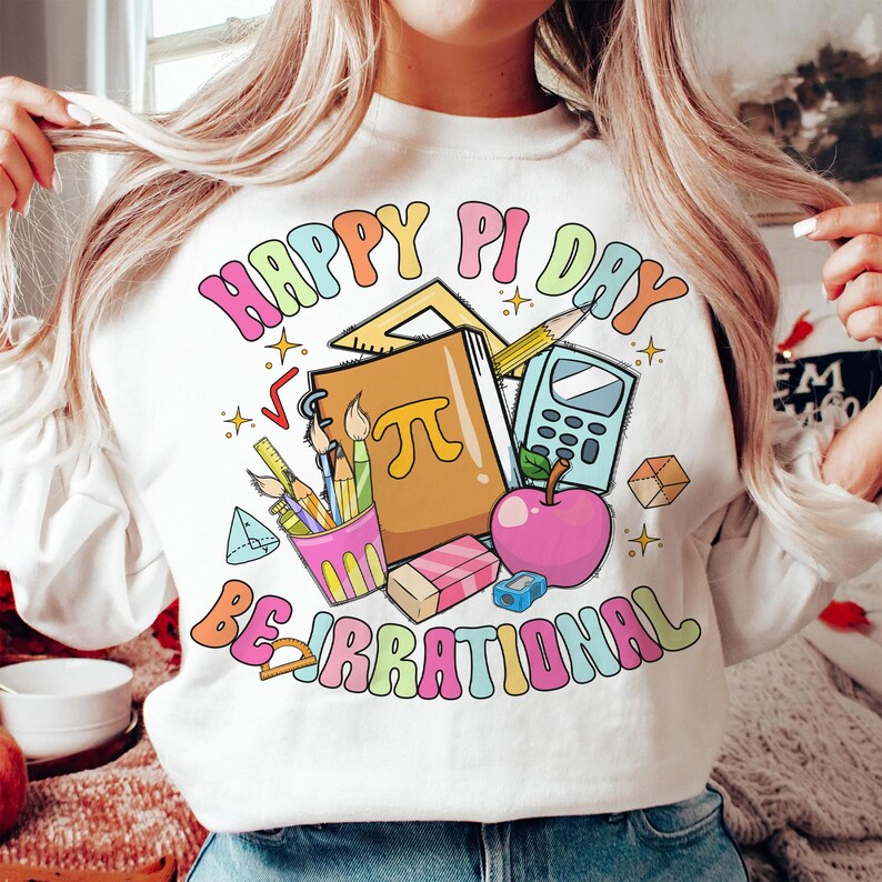 May include: White sweatshirt with a colorful graphic design that says "Happy Pi Day Be Irrational". The graphic includes a notebook, pencils, a calculator, a protractor, and a red apple.