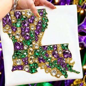 May include: A white square canvas featuring a jeweled outline of the state of Louisiana. The design is filled with purple, green, and clear gemstones, with gold-colored borders. The gemstones are arranged to resemble the state's shape.