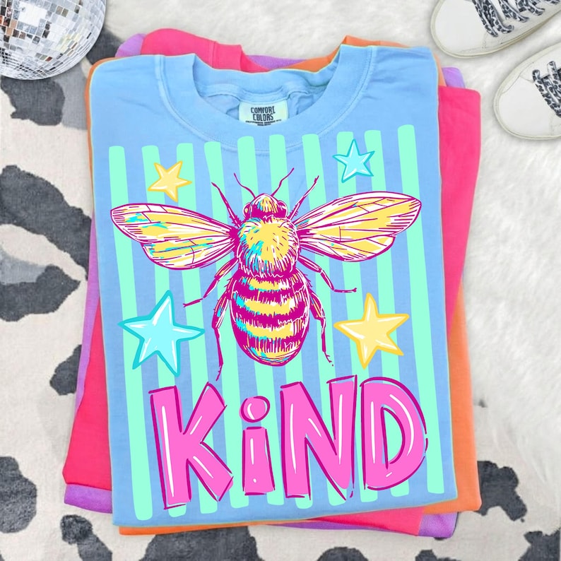 Puede incluir: Una camiseta Comfort Colors azul claro con un gr&aacute;fico de abeja colorida, rayas verticales y estrellas. La palabra "KIND" est&aacute; impresa en rosa. La camiseta est&aacute; doblada encima de otras camisetas coloridas.