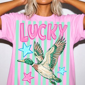 Puede incluir: Camiseta rosa con la palabra "LUCKY" en letras rosas con burbujas. La camiseta presenta un gr&aacute;fico de un pato volador con plumas verdes, marrones y blancas, y estrellas azules y rosas. El fondo tiene rayas verticales rosas y verdes.