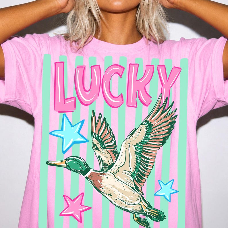 Puede incluir: Camiseta rosa con la palabra "LUCKY" en letras rosas con burbujas. La camiseta presenta un gr&aacute;fico de un pato volador con plumas verdes, marrones y blancas, y estrellas azules y rosas. El fondo tiene rayas verticales rosas y verdes.