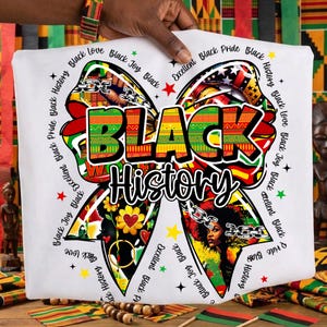 May include: White t-shirt featuring a large bow design with the words "BLACK HISTORY" in bold, colorful letters. The bow is decorated with various patterns, images of a woman, and the phrases "Black Love," "Black Joy," and "Black Pride."