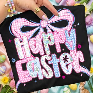 May include: Black t-shirt featuring a pink and blue bow and the words "Happy Easter". The letters are decorated with floral, polka dot, and gingham patterns. A festive Easter-themed graphic tee.