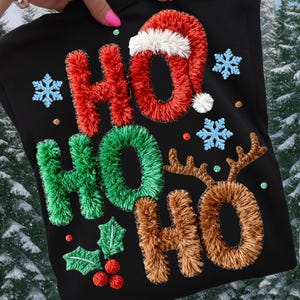 Puede incluir: Sudadera negra con diseño navideño bordado. Presenta las palabras "HO HO HO" en rojo, verde y marrón, con un gorro de Papá Noel, copos de nieve, acebo y cuernos de reno. Ropa festiva.