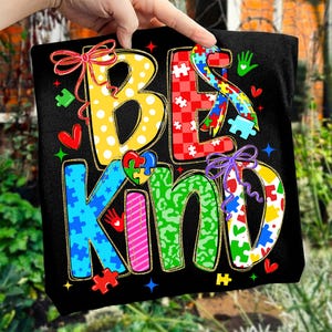 May include: Black t-shirt with the words "BE KIND" in colorful, patterned letters. The letters are decorated with polka dots, puzzle pieces, hearts, and bows. The design has a gold outline.