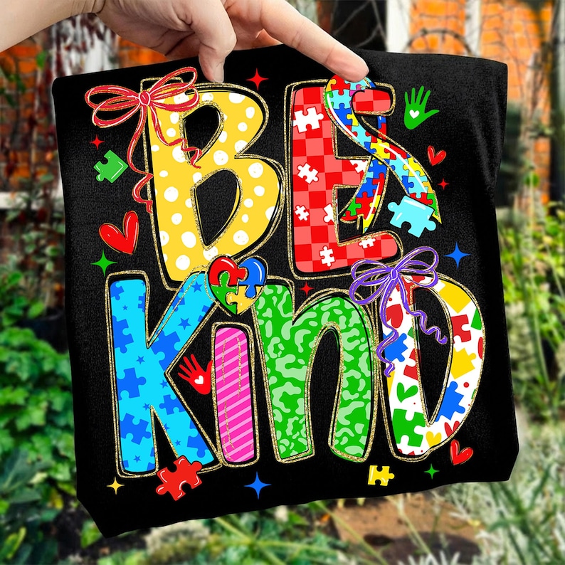 May include: Black t-shirt with the words "BE KIND" in colorful, patterned letters. The letters are decorated with polka dots, puzzle pieces, hearts, and bows. The design has a gold outline.