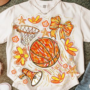 May include: Cream-colored t-shirt featuring a basketball design with orange, yellow, and gold accents. The graphic includes a basketball, flowers, stars, a bow, a megaphone, and a basketball hoop. The shirt has the text "Comfort Colors" on the collar.