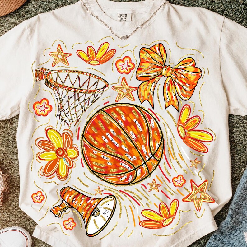 May include: Cream-colored t-shirt featuring a basketball design with orange, yellow, and gold accents. The graphic includes a basketball, flowers, stars, a bow, a megaphone, and a basketball hoop. The shirt has the text "Comfort Colors" on the collar.