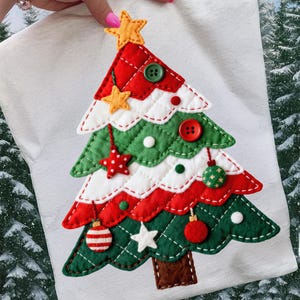 Könnte beinhalten: Ein weißes, weihnachtsbaumförmiges Applikationsmuster mit rot, grün und weiß gesteppten Abschnitten. Der Baum ist mit Stern- und Kugelornamenten, Knöpfen und einem Stern als Spitze verziert. Der Baum ist etwa 25 cm hoch.