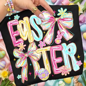 May include: Black t-shirt with the word "EASTER" in large, colorful letters. The letters are outlined in white and filled with yellow, pink, and blue. The design includes bows, flowers, and an Easter egg.