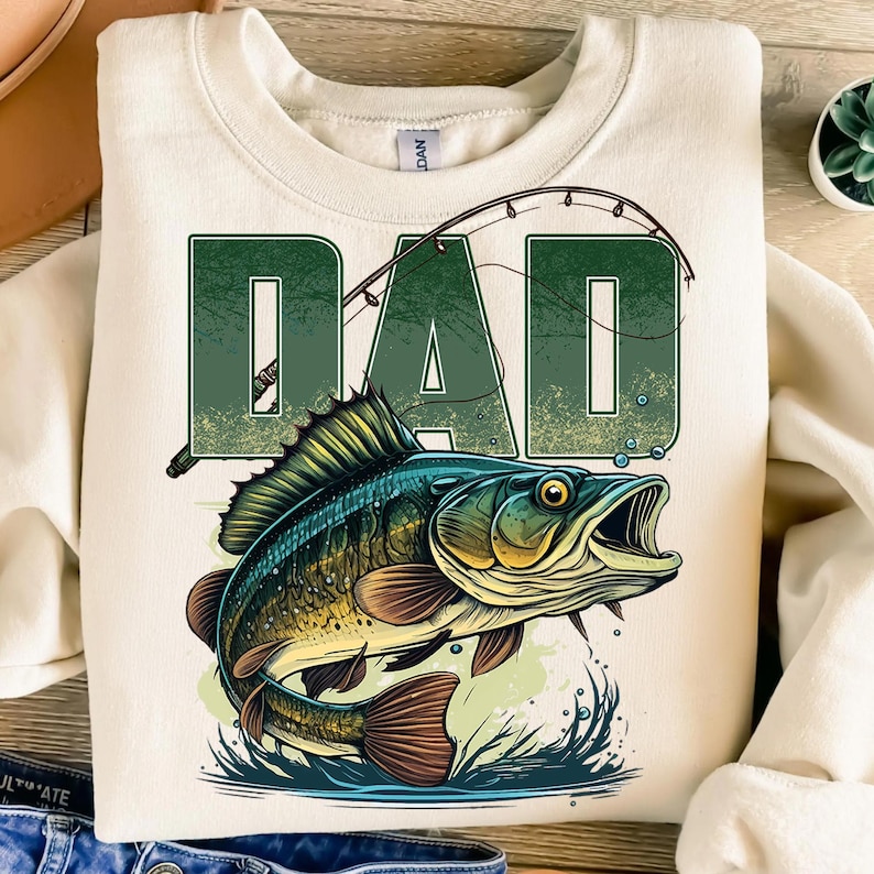 Pu&ograve; includere: Felpa color crema con grafica verde "DAD" e design a canna da pesca. Sotto il testo, un'illustrazione dettagliata di un pesce che salta fuori dall'acqua, con colori blu, verdi e marroni.