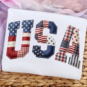 May include: White t-shirt with the word "USA" embroidered in red, white, and blue. The letters are designed with patchwork patterns and the American flag, perfect for patriotic celebrations.