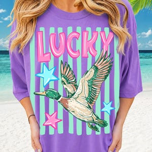 Puede incluir: Una camiseta morada con un gr&aacute;fico de un pato volador, estrellas y la palabra "LUCKY" en rosa y blanco. La camiseta tiene rayas verticales de color verde menta. El pato tiene detalles verdes, marrones y blancos.