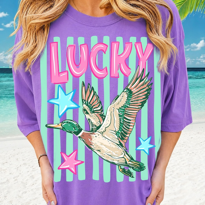 Puede incluir: Una camiseta morada con un gr&aacute;fico de un pato volador, estrellas y la palabra "LUCKY" en rosa y blanco. La camiseta tiene rayas verticales de color verde menta. El pato tiene detalles verdes, marrones y blancos.