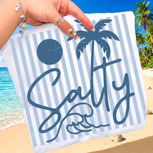 Puede incluir: Una toalla de playa blanca con rayas azules y la palabra "Salty" en escritura cursiva. El diseño incluye una palmera azul, un sol y olas. La toalla se sostiene en una playa de arena con palmeras al fondo.
