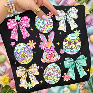 May include: Black t-shirt with a vibrant Easter design. The print showcases decorated Easter eggs, bows, and a bunny with a heart. The artwork uses bright pastel colours with floral and polka dot details, ideal for the Easter season.