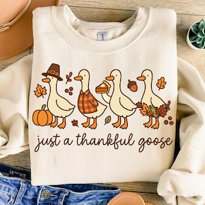 May include: Cream-colored sweatshirt with a graphic of four cartoon geese in fall attire. The geese are wearing a hat, plaid shirt, and holding a pie and an acorn. The text "just a thankful goose" is below the geese.