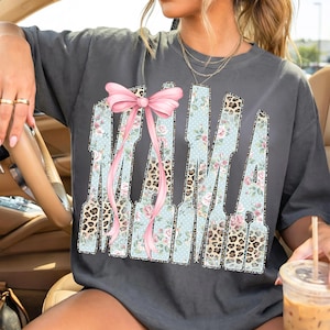May include: Dark gray t-shirt with the word "MAMA" in a unique design. The letters feature floral, leopard print, and polka dot patterns, with a pink bow accent. A casual, stylish top.