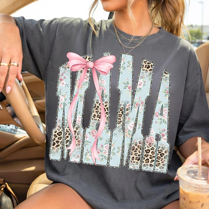 May include: Dark gray t-shirt with the word "MAMA" in a unique design. The letters feature floral, leopard print, and polka dot patterns, with a pink bow accent. A casual, stylish top.