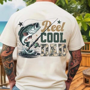 Może przedstawiać: Kremowa koszulka z grafiką przedstawiającą rybę wyskakującą z wody, wędkę i napisem "Reel Cool Dad". Tekst jest w kolorze brązowym, zielonym i moro. Projekt zawiera gwiazdy i pluski wody.