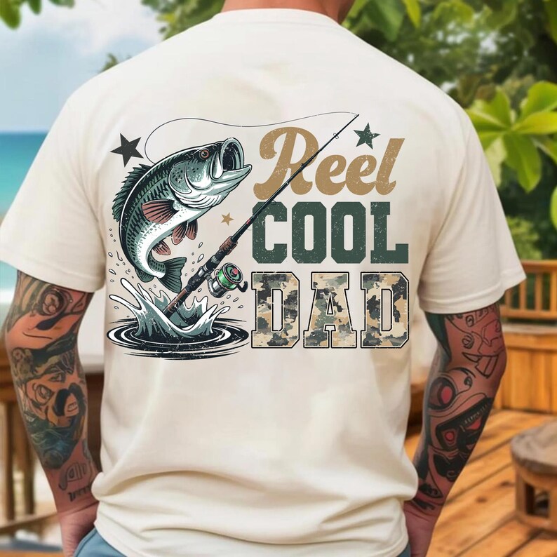 Może przedstawiać: Kremowa koszulka z grafiką przedstawiającą rybę wyskakującą z wody, wędkę i napisem "Reel Cool Dad". Tekst jest w kolorze brązowym, zielonym i moro. Projekt zawiera gwiazdy i pluski wody.