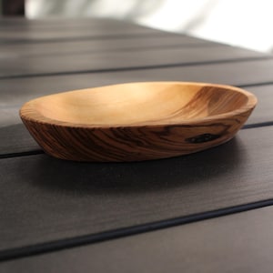 Könnte beinhalten: Eine handgefertigte Holzschale mit einem natürlichen, hellen Innenraum und einem dunkleren, gestreiften Äußeren. Die ovale Schale steht auf einer dunklen Oberfläche und zeigt die Holzmaserung und Handwerkskunst. Ideal zum Servieren oder Ausstellen.