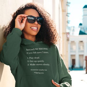 Puede incluir: Una sudadera con capucha verde oscuro con el texto "Secret to success: If you fall down 7 times, 1. Play dead. 2. Get up quietly. 3. Make moves silently. Someone nearby was tripping you." La sudadera con capucha la lleva una persona con gafas de sol.