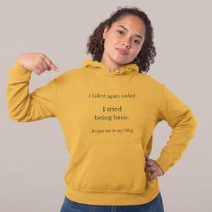 Op de afbeelding: Mosterdgele hoodie met een voorzak en een capuchon met trekkoord. De tekst op de voorkant luidt: "I failed again today. I tried being basic. It's just not in my DNA."
