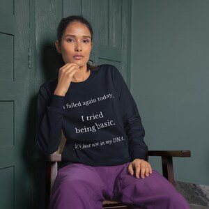 Könnte beinhalten: Ein marineblaues Sweatshirt mit weißem Text: "Ich habe heute wieder versagt. Ich habe versucht, einfach zu sein. Es liegt einfach nicht in meiner DNA." Die Person trägt eine lila Hose und sitzt auf einem Holzstuhl.