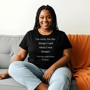 May include: A black t-shirt with the text "I'm sorry for the things I said when I was hungry. Next time, bring snacks. Do better." The shirt is worn by a person sitting on a gray couch with an orange blanket.