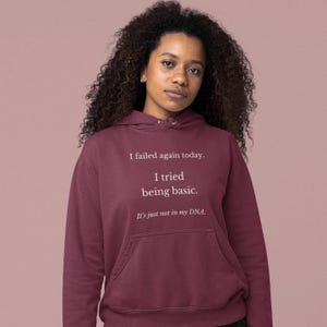 Op de afbeelding: Een bordeauxrode hoodie met de tekst "I failed again today. I tried being basic. It's just not in my DNA." De hoodie heeft een voorzak en een capuchon. De persoon op de foto draagt de hoodie.