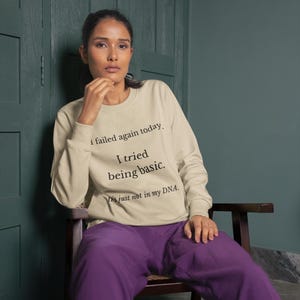 Op de afbeelding: Een beige sweatshirt met de tekst "I failed again today. I tried being basic. It's just not in my DNA." De persoon draagt een paarse broek en zit op een houten stoel.