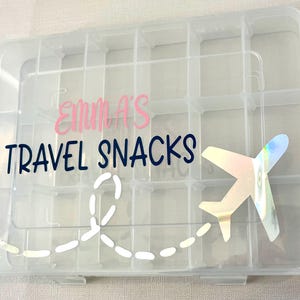 May include: A clear plastic storage box with a lid, divided into 16 compartments. The box has a pink and blue sticker that says "Emma's Travel Snacks" with a white airplane and a dotted line trail.