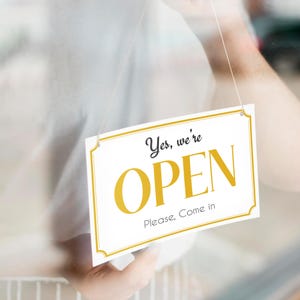 May include: A white sign with a gold border and gold text that reads "Yes, we're OPEN. Please, come in." The sign is hanging from a string.