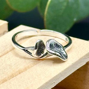 May include: A silver ring featuring a leaf design. The ring has two detailed leaf shapes, with visible veins, connected to the band. The ring is set against a light wooden surface, with a blurred green background.