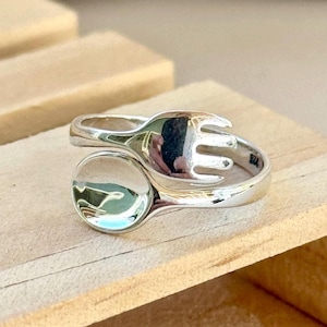 May include: A silver ring with a fork and spoon design. The ring features a fork on one end and a spoon-shaped design on the other, with a smooth, polished finish. The ring is set against a light wooden surface.