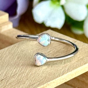 May include: A silver-colored open ring with two heart-shaped opal stones. The ring is set against a wooden surface, with white flowers and purple petals in the background. The opal stones have a pearly, iridescent appearance.