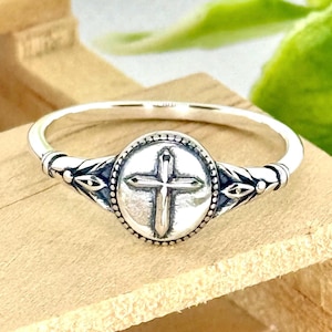 May include: A silver ring featuring a raised cross design within a circular setting. The ring has a thin band and decorative accents on either side of the cross. The ring is displayed on a wooden surface.