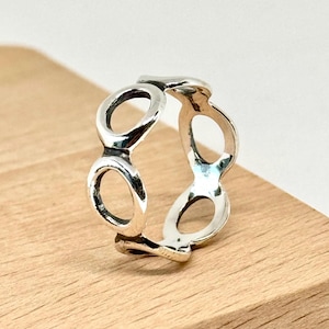 May include: A silver ring featuring a repeating open-circle design. The ring is made of a shiny, polished metal and sits on a wooden surface. The design is continuous, with each circle connected to the next, creating a unique pattern.