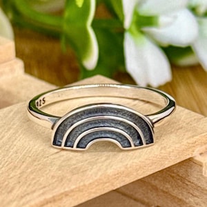 May include: A silver ring with a rainbow design. The ring features a detailed, arched rainbow with multiple layers. The band is smooth and shiny. The number "125" is engraved on the ring. The ring is set on a wooden surface.