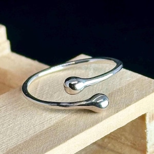 May include: A silver-colored ring with a unique design. The ring features a slender band that curves around and ends with two rounded, teardrop-shaped accents. The ring is displayed on a light-colored wooden surface.