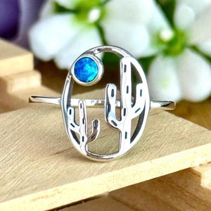 May include: A silver ring featuring a desert cactus design. The ring has a central oval with two cactus silhouettes and a blue gemstone. The ring is set on a wooden surface with a blurred floral background.