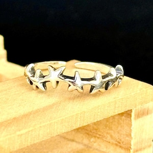 May include: A silver-colored ring featuring a series of star-shaped designs. The ring is set against a wooden surface with a dark background. The stars are evenly spaced and appear to be connected, creating a celestial theme.