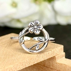 May include: A silver-toned ring featuring a floral design. The ring has a circular band with a flower at the top and leaf accents. The ring is set on a wooden surface.