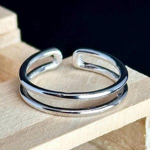 May include: A silver-colored ring with a double-band design. The ring has a smooth, polished finish and an open design at the top. The ring is displayed on a light-colored wooden surface.