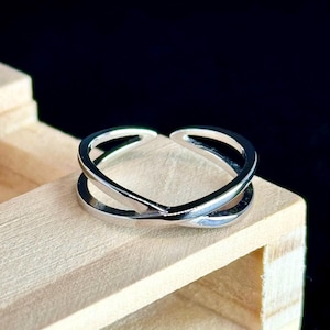 May include: A silver-colored ring with a crisscross design. The ring is made of a shiny metal and sits on a light-colored wooden surface. The ring's design features two bands that intersect, creating an open, modern look.