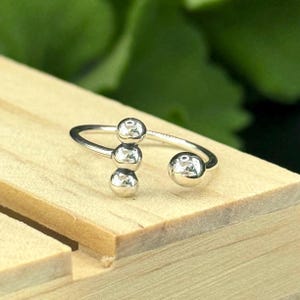 May include: A silver-colored ring with a unique design. The ring features three small, round beads clustered together, and a larger bead on the opposite end. The ring is open and adjustable.