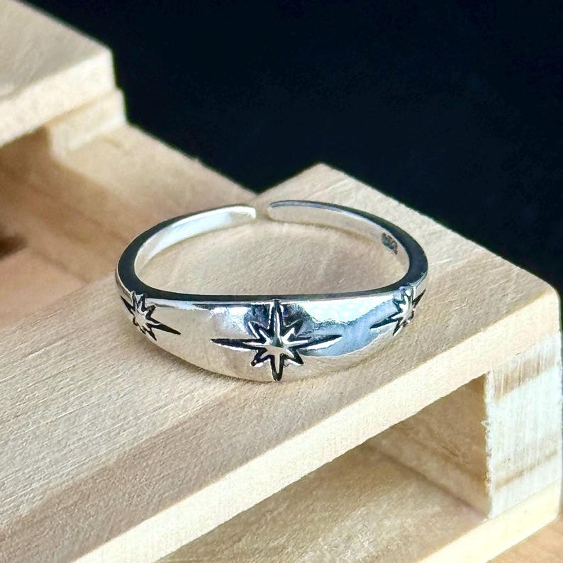 May include: A silver-colored ring with a starburst design. The ring features a central starburst and smaller starbursts on either side. The ring is set against a light wooden surface.