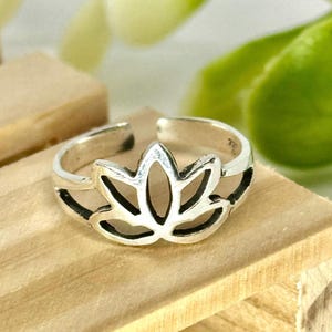 May include: A silver-colored toe ring featuring a lotus flower design. The ring has an open design, allowing for adjustability. The lotus flower is the focal point, with detailed cut-out petals. The ring sits on a wooden surface.
